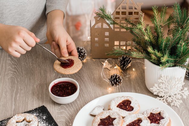 As mãos das crianças espalham geléia de baga em biscoitos Linzer na mesa da cozinha. Cozinhar guloseimas de Natal. Estilo de vida