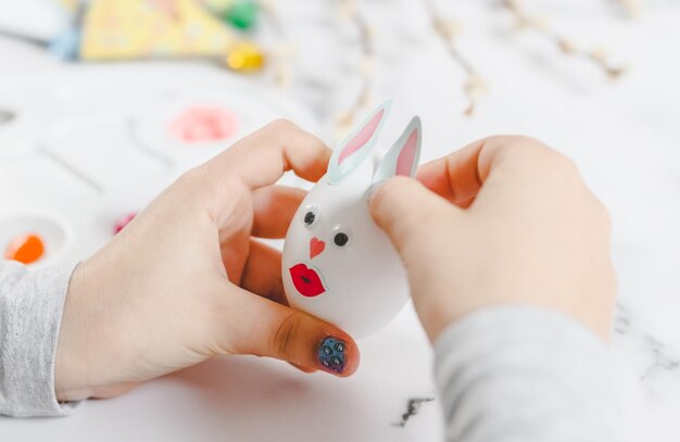 As mãos das crianças seguram um ovo branco com olhos colados, lábios e orelhas de coelho de papel