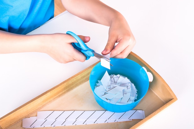 As mãos das crianças seguram uma tesoura azul e cortam o papel.