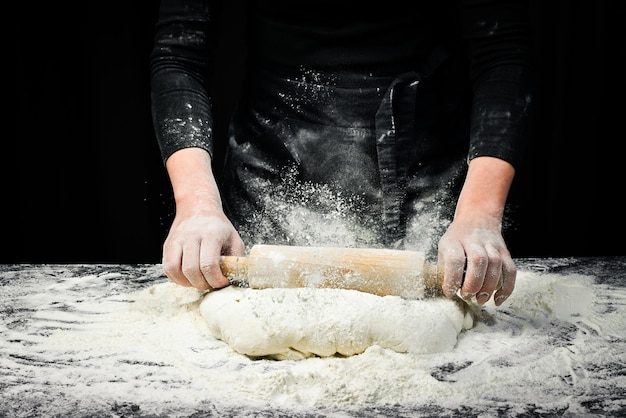 As mãos das mulheres com um rolo preparam a massa para pão ou pizza Fundo de cozimento preto