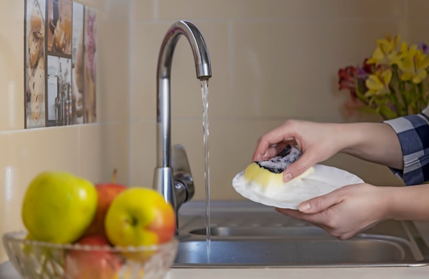 As mãos das mulheres lavam um prato branco com uma esponja sob água corrente na pia da cozinha Lavando pratos em casa