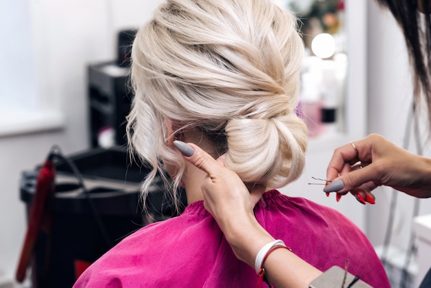 As mãos de um cabeleireiro profissional fazem um penteado de coque baixo para uma menina loira