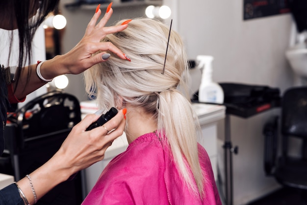 As mãos de um cabeleireiro profissional fazem um penteado de coque baixo para uma menina loira