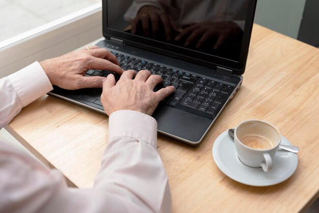 As mãos de um homem mais velho irreconhecível digitando no teclado de um laptop uma xícara de café na mesa