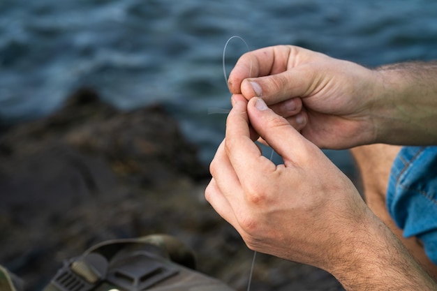 As mãos de um pescador pegando a linha para dar um nó ao fundo uma mochila em algumas pedras