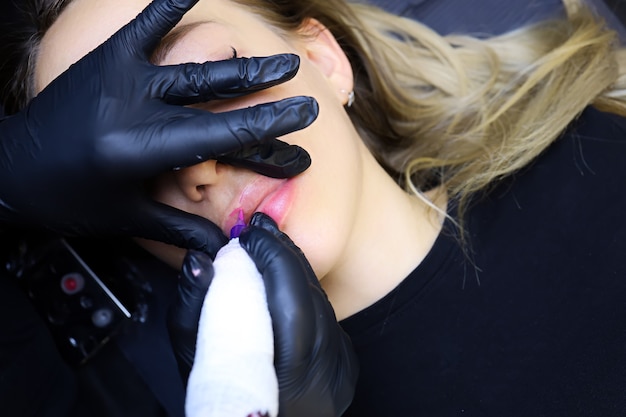 As mãos de um tatuador em luvas pretas seguram os lábios da modelo e fazem a maquiagem definitiva dos lábios com uma máquina de tatuagem