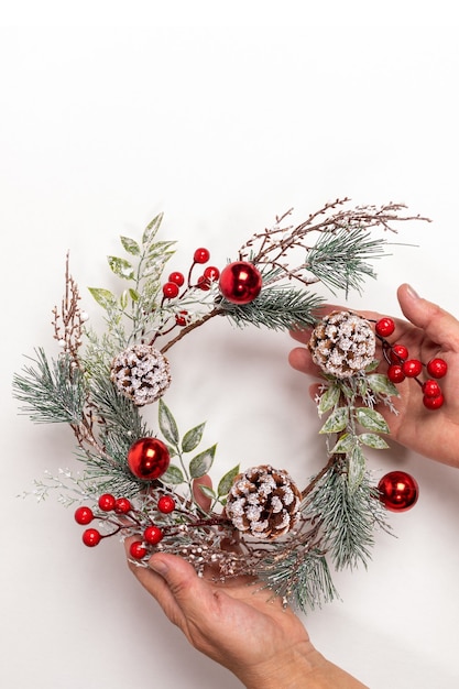 As mãos de uma mulher estão segurando uma guirlanda de natal reutilizável feita à mão, feita de galhos de árvores, frutas vermelhas, cones em branco