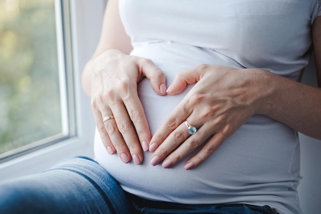 As mãos de uma mulher, formando um símbolo de coração em uma barriga de grávida. Uma mulher grávida de camiseta branca está sentada perto da janela.