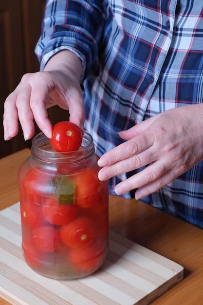 As mãos de uma mulher idosa tiram deliciosos tomates em conserva caseiros de uma jarra na cozinha em uma mesa marrom sem rosto fechado