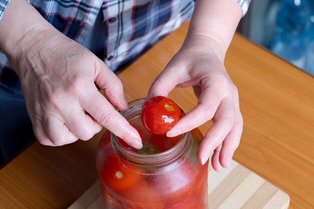 As mãos de uma mulher idosa tiram deliciosos tomates em conserva caseiros de uma jarra na cozinha em uma mesa marrom sem rosto fechado