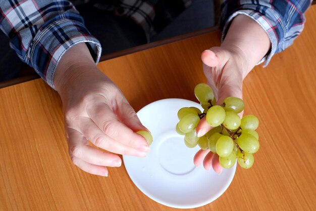 As mãos de uma senhora idosa seguram um pequeno pincel de uvas brancas sem sementes e as exibem na cozinha em uma mesa marrom sem rosto fechado