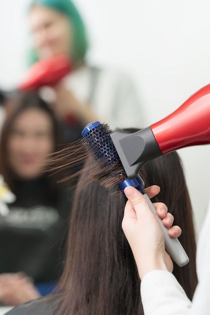 As mãos do cabeleireiro secam os cabelos morenos da cliente, usando o secador de cabelo vermelho e o pente azul no salão de beleza profissional.