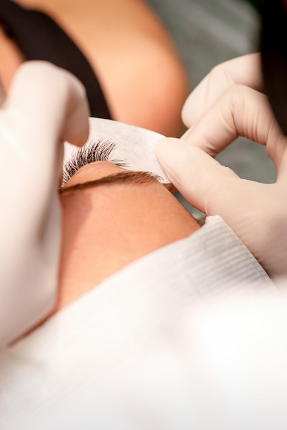 As mãos do cosmetologista estão colando fita branca sob o olho da jovem mulher caucasiana durante o procedimento de extensão dos cílios closeup