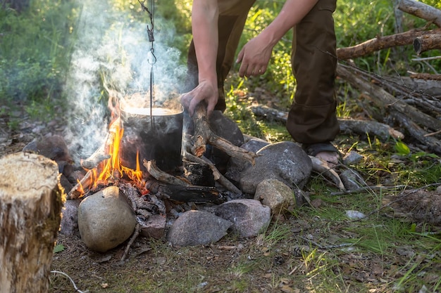 As mãos do homem estão colocando lenha na fogueira sobre a qual pendura uma panela com um jantar preparando