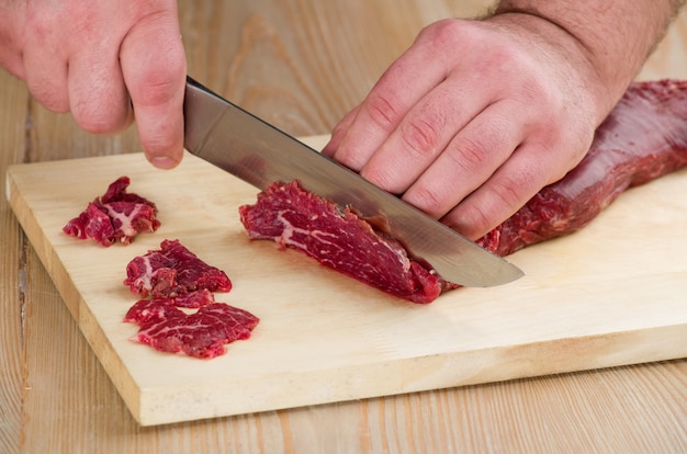 As mãos do homem estão cortando carne crua por uma faca em uma mesa de madeira