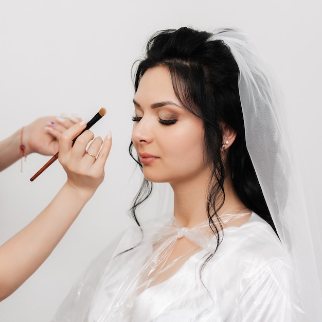 As mãos do maquiador com pincel maquia e aplica sombras nos olhos da noiva no salão de beleza