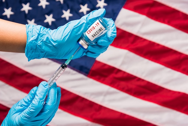As mãos do médico usam luvas segurando uma seringa e um frasco de vacina COVID de coronavírus na bandeira dos Estados Unidos da América