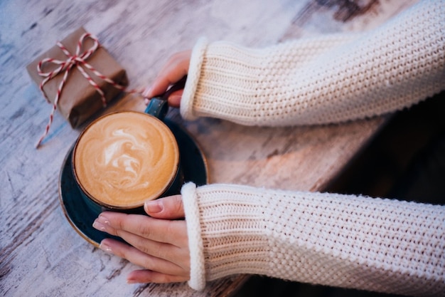 As mãos femininas estão segurando uma xícara de café. A atmosfera de uma noite acolhedora. Bebida quente. Uma garota com um suéter branco quente. presentes de natal e ano novo.