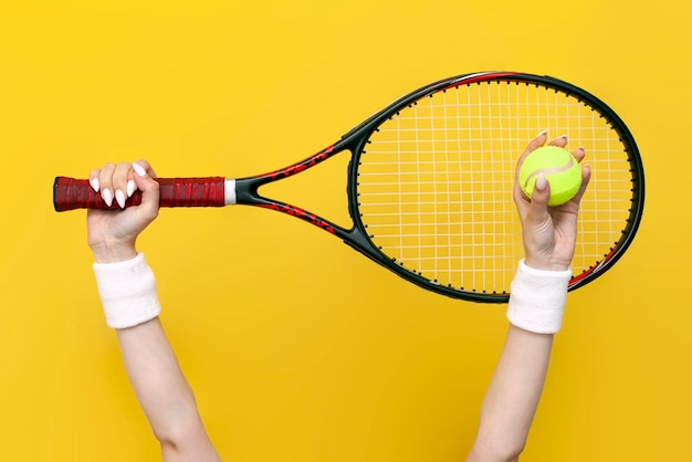 As mãos femininas levantam e seguram a raquete de tênis e a bola de tênis em fundo amarelo isolado