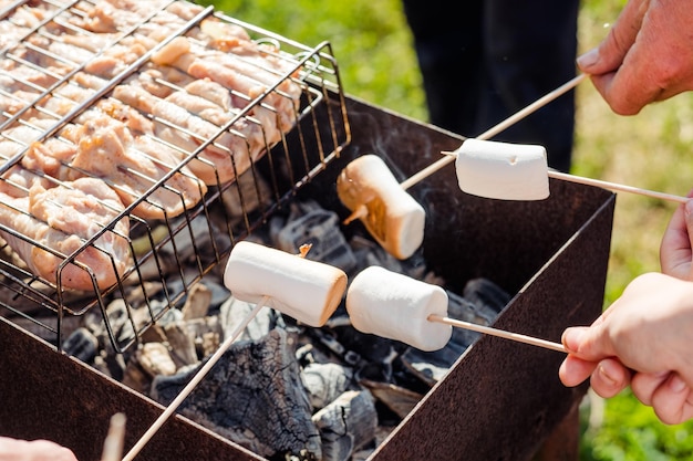 As mãos seguram marshmallows em um espeto de madeira sobre a grelha marshmallows fritos e carne grelhada