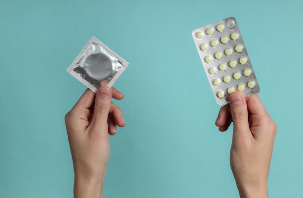 As mãos seguram preservativos na embalagem, pílulas anticoncepcionais em papel azul