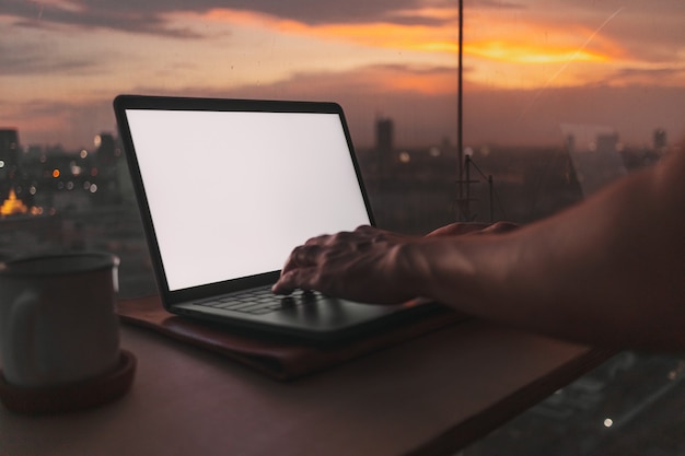 As mãos trabalham no laptop de tela vazia com vista da cidade ao pôr do sol à noite