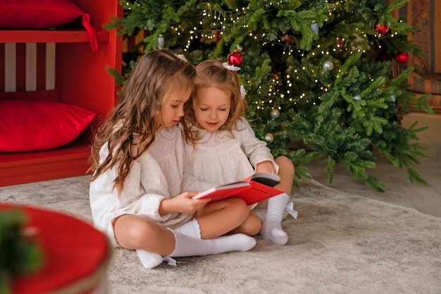 As meninas estão sentadas no chão perto de uma árvore de Natal e lendo um livro