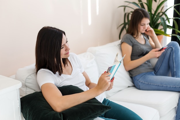 As meninas estão sentadas no sofá olhando para o telefone e o livro.