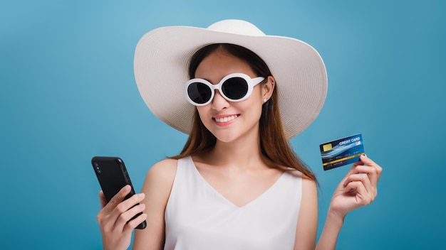 As mulheres bonitas sorridentes asiáticas usam um chapéu largo e óculos de sol usando smartphones compras on-line com cartão de crédito isolado em fundo de cor azul Conceito de negócios de viagens com promoção e venda