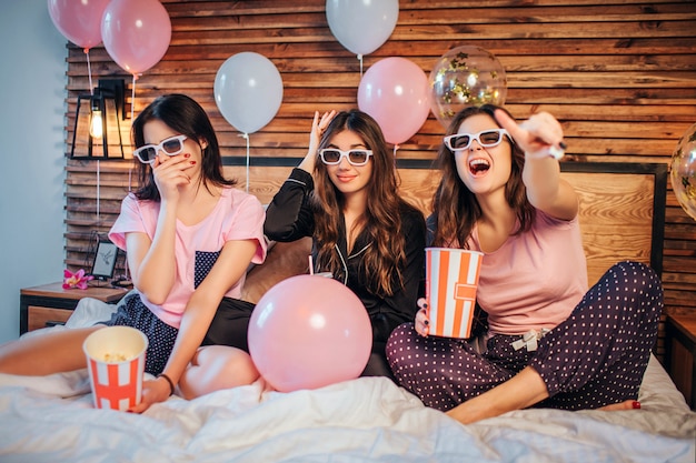 As mulheres jovens emocionais sentam-se na cama na sala festiva. Eles assistem filmes através de óculos especiais. as mulheres usam pijamas. Eles têm festa.