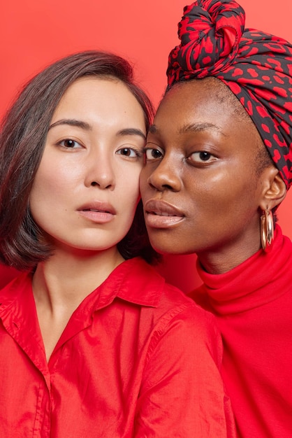 As mulheres têm cores diferentes de aparência específica da pele, ficam próximas umas das outras, olham com confiança para a câmera, usam roupas elegantes isoladas no vermelho, sendo melhores amigas