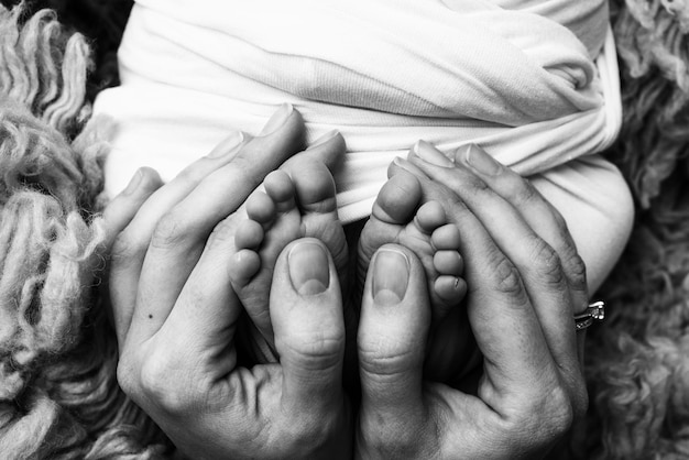 As palmas das mãos do pai, a mãe estão segurando o pé do bebê recém-nascido. Pés do recém-nascido nas palmas dos pais. Fotografia de estúdio dos dedos dos pés, saltos e pés de uma criança. Preto branco.