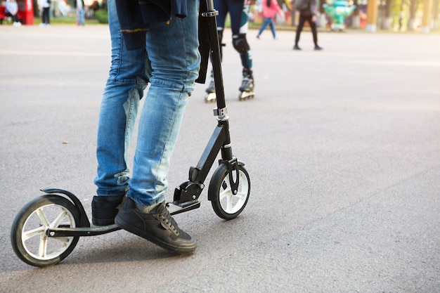 As pernas de um homem de jeans e tênis em uma scooter no parque em uma pista de asfalto. Caminhada esportiva, estilo de vida ativo e saudável, transporte ecologicamente correto, trânsito