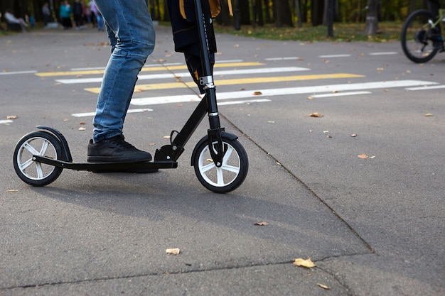 As pernas de um homem de jeans e tênis em uma scooter no parque no outono com folhas amarelas secas caídas no asfalto. Caminhadas no outono, estilo de vida ativo, transporte ecológico, trânsito