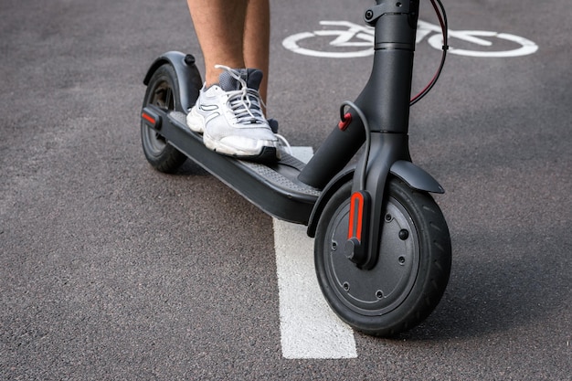 As pernas do homem de tênis branco andam em uma scooter elétrica preta sobre asfalto