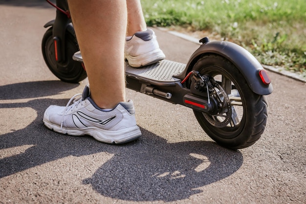 As pernas do homem de tênis branco andam em uma scooter elétrica preta sobre asfalto