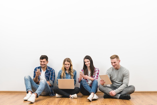 As quatro pessoas felizes sentadas no chão com gadgets no fundo branco