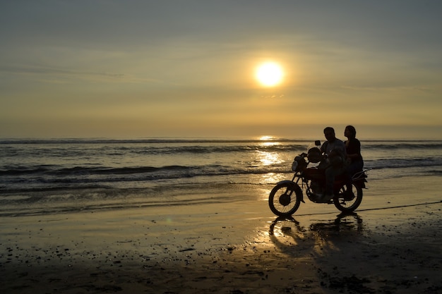 As silhuetas escuras de um casal em uma motocicleta caminhando pela praia ao pôr do sol