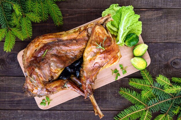 Asar conejo entero relleno de ciruelas pasas sobre una tabla de madera con zanahorias al horno y coles de Bruselas