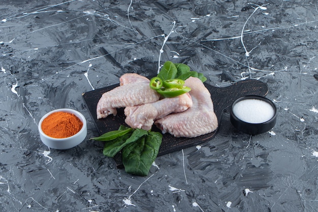 Asas de frango cru e espinafre em uma placa de corte ao lado de uma tigela de especiarias e sal, na superfície de mármore.