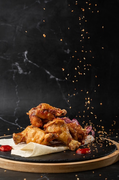 Asas de frango de búfalo assado com sementes de gergelim, cebola e fatias de pimentão na placa de madeira preta.