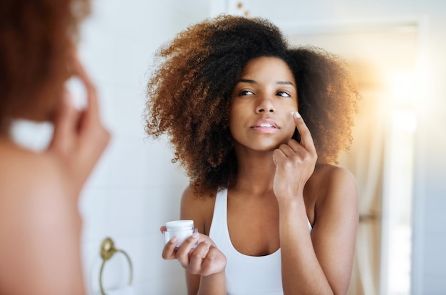 Asegurándose de que su piel permanezca hidratada todo el día Foto de una mujer joven y atractiva que se aplica crema hidratante en la cara frente al espejo