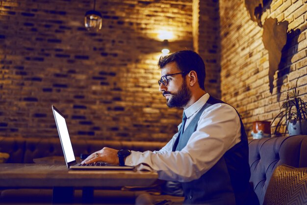 Asesor financiero barbudo caucásico sofisticado serio en traje con anteojos sentado en la cafetería y usando la computadora portátil.