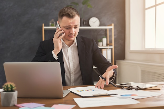 Asesor financiero masculino serio que trabaja con papeles y consultoría en dispositivos móviles. Hombre en ropa formal revisando documentos y hablando por teléfono inteligente en el interior de la oficina moderna, copie el espacio en la pared negra