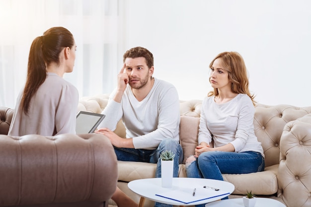 Asesoramiento psicológico. Triste infeliz pareja joven mirando a la psicóloga y escuchándola mientras está sentado en el sofá