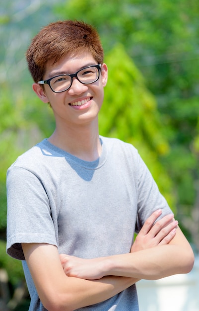 Foto asia adolescente guapo con gafas de pie con los brazos cruzados, retrato alegre hipster joven está mirando y sonríe con una cara feliz en el parque en tailandia