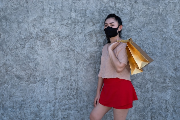 Asia joven mujer vistiendo una mascarilla con la celebración de bolsas de la compra en el fondo de la pared de hormigón