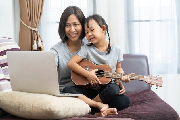 Asia madre e hija usando laptop y ukelele en casa