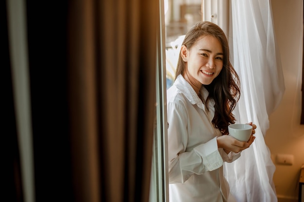 Asia mujer joven que vive en casa relajarse y beber una taza de café caliente en el dormitorio de vacaciones. Asia, asia, relajarse, solo, tecnología, concepto de estilo de vida.