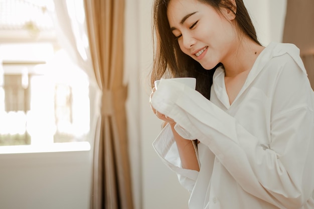 Asia mujer joven que vive en casa relajarse y beber una taza de café caliente en el dormitorio de vacaciones. Asia, asia, relajarse, solo, tecnología, concepto de estilo de vida.